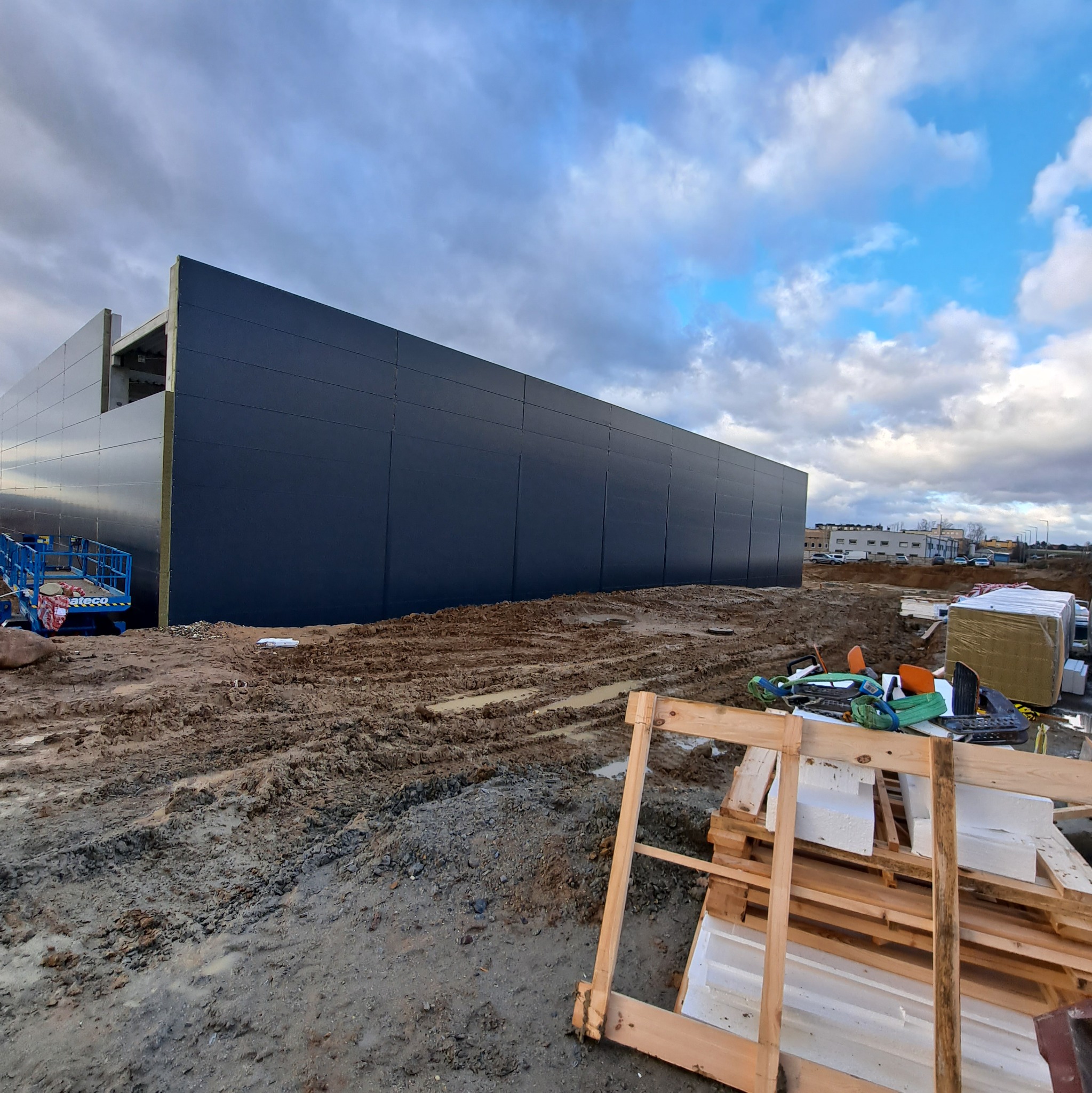 Budynek w trakcie budowy z szarymi płytami warstwowymi na ścianach, widoczny podnośnik koszowy i materiały budowlane na błotnistym placu budowy pod pochmurnym niebem.
