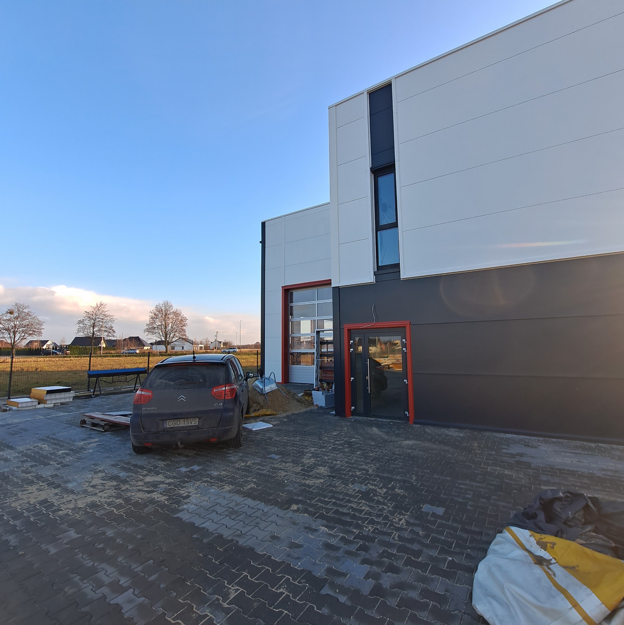 Nowoczesny budynek przemysłowy z płyt warstwowych w kolorach białym i grafitowym, z czerwoną stolarką okienną i drzwiową, częściowo ukończony teren wokół budynku, zaparkowany samochód osobowy.