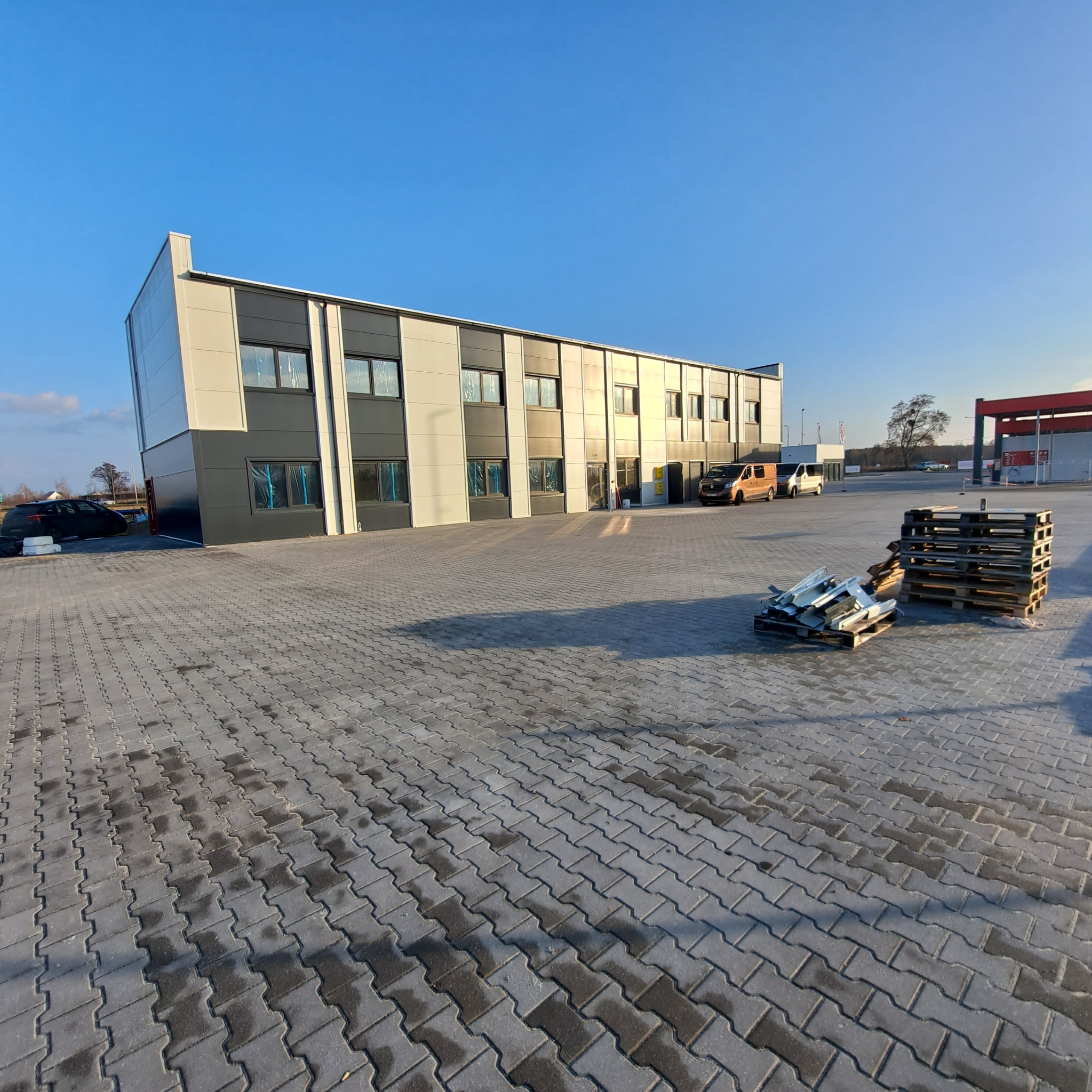 Nowoczesny budynek biurowy z płyt warstwowych w kolorach szarości i bieli, widok z zewnątrz, palety i resztki materiałów budowlanych na pierwszym planie.