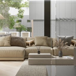 Sayna Polygons - Beżowa sofa z poduszkami w salonie z dużymi oknami i widokiem na zieleń. Na stoliku wazon z suszonymi kwiatami i książka 'Bauhaus'.