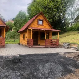 Dwa drewniane domki letniskowe z czerwonymi dachami i werandami, nowo ułożona szara kostka brukowa, czarna podsypka pod kostkę, tło z zielenią drzew.