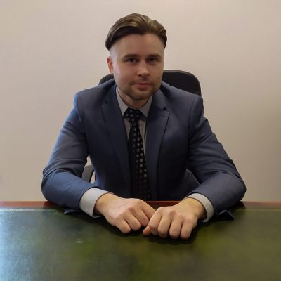 Portret adwokata w niebieskim garniturze siedzącego za biurkiem z zielonym blatem, dłonie splecione na biurku.