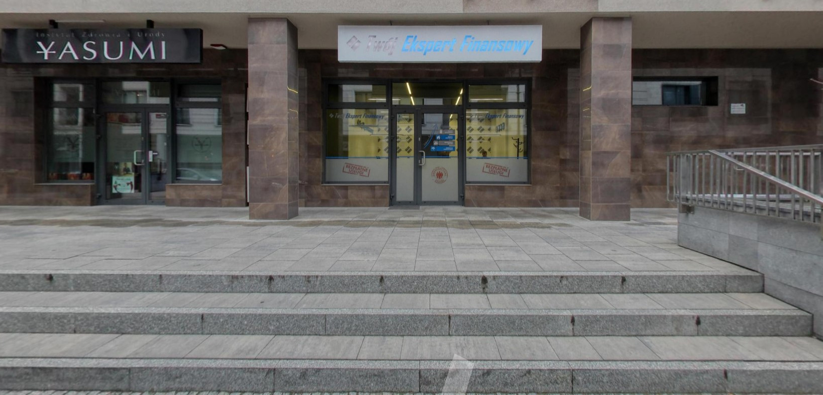 Wejście do biura 'Twój Ekspert Finansowy' z widoczną tablicą informacyjną i naklejkami na drzwiach o bezpłatnych usługach, sfotografowane z dołu schodów.