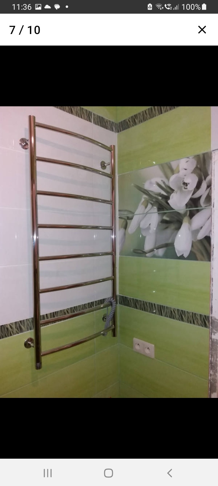 Chromowany grzejnik drabinkowy elektryczny zamontowany na ścianie w łazience z zielonymi płytkami i dekoracyjnym panelem z motywem kwiatowym.