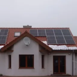 Dom jednorodzinny z panelami fotowoltaicznymi na czerwonym dachu pokrytym śniegiem, widoczna antena satelitarna na elewacji.