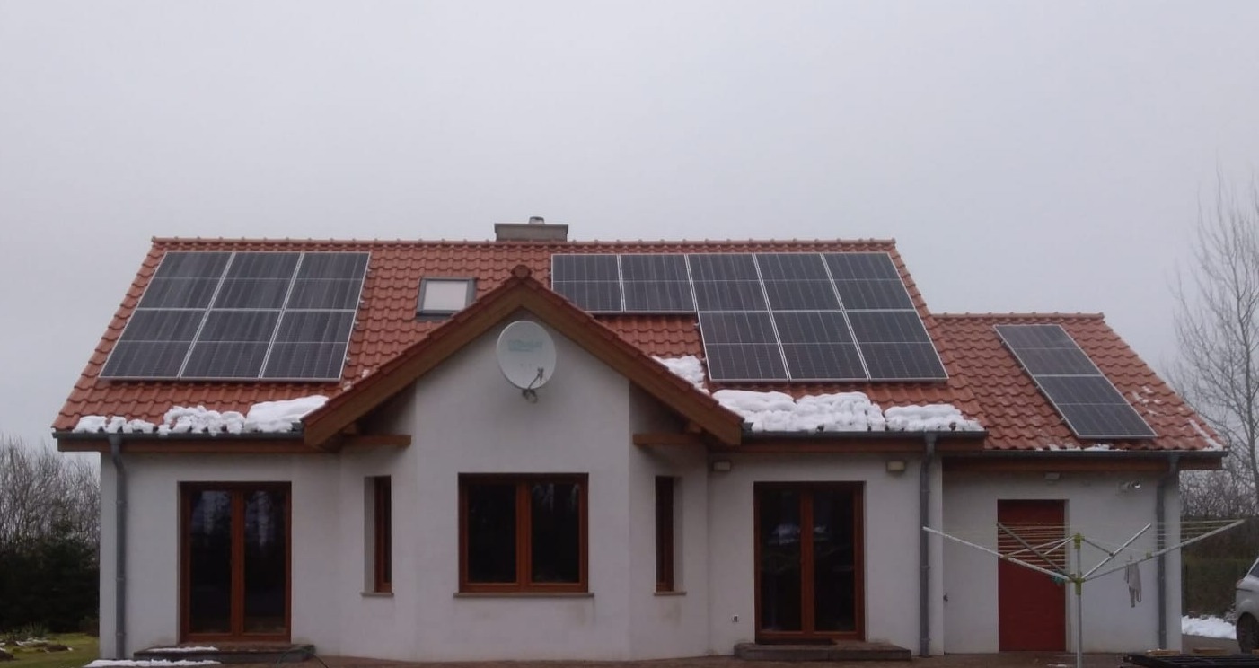 Dom jednorodzinny z panelami fotowoltaicznymi na czerwonym dachu pokrytym śniegiem, widoczna antena satelitarna na elewacji.