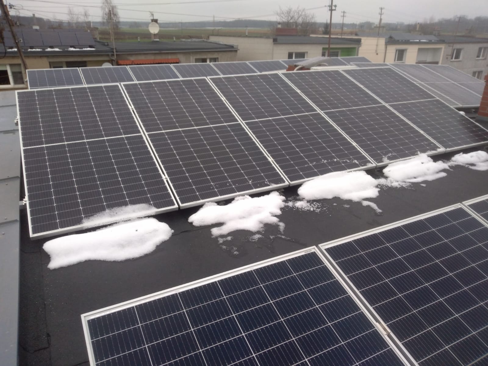 Panele słoneczne na dachu budynku częściowo pokryte topniejącym śniegiem w pochmurny dzień.