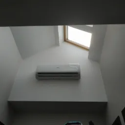 Biały klimatyzator Haier zamontowany na skośnej ścianie pod oknem dachowym, widok z dołu.