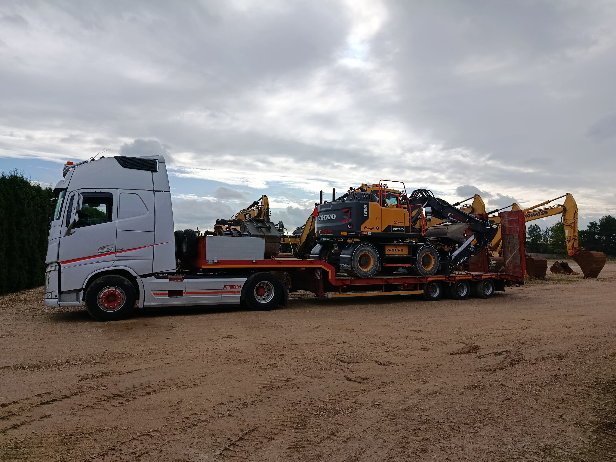 Ciężarówka transportuje pomarańczową koparkę Volvo na niskopodwoziowej platformie. W tle widoczne inne maszyny budowlane i pochmurne niebo.