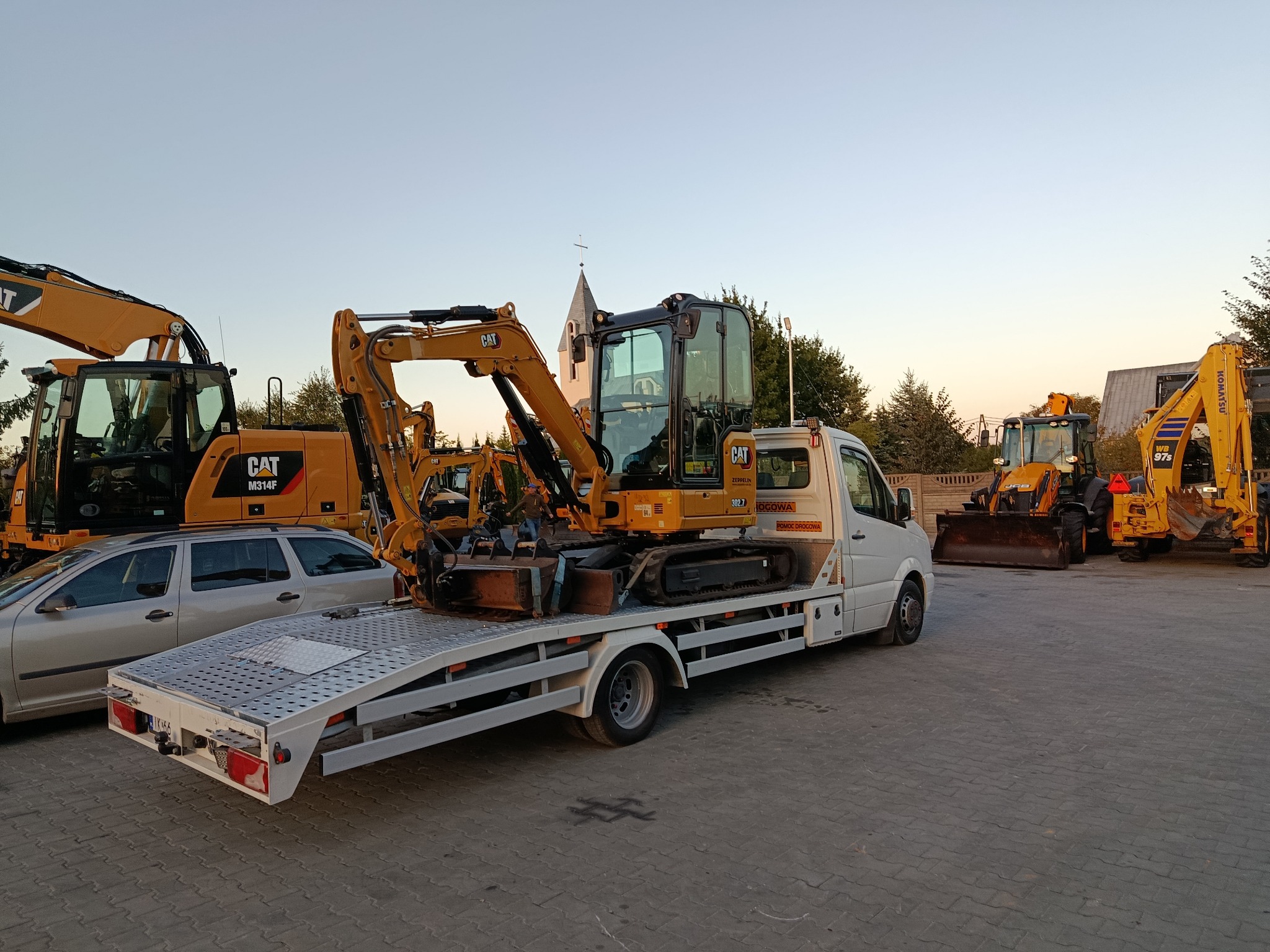Laweta z mini koparką CAT na platformie, w tle inne maszyny budowlane i kościół. Pomoc drogowa dla maszyn budowlanych.