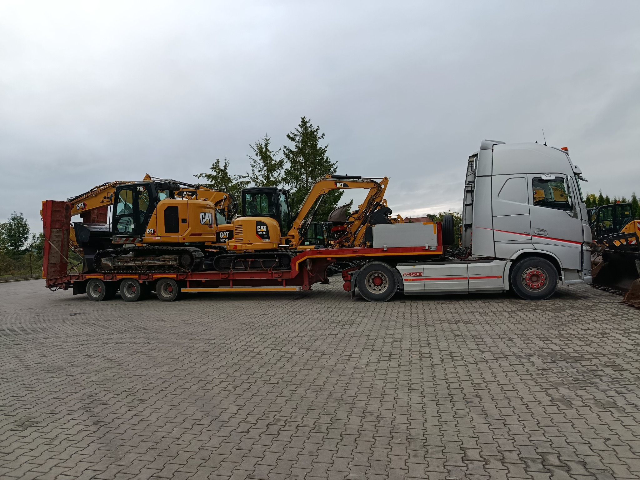 Laweta przewozi dwie żółte koparki CAT na niskopodwoziowej przyczepie. Szary ciągnik siodłowy Volvo FH500. Transport ciężkiego sprzętu budowlanego na placu.