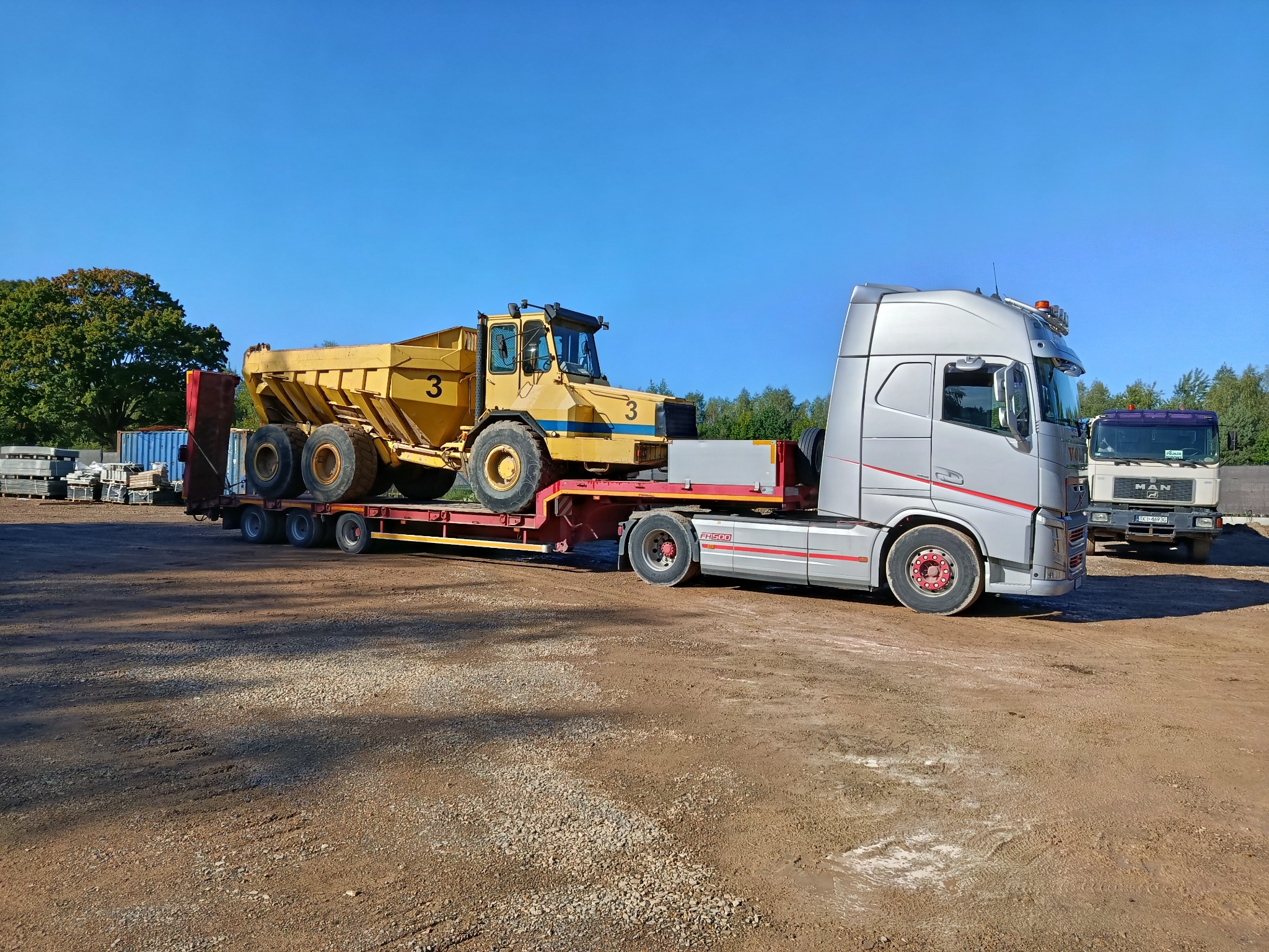 Żółta wywrotka budowlana na niskopodwoziowej platformie transportowej, ciągniętej przez srebrną ciężarówkę Volvo FH500, na żwirowym placu.