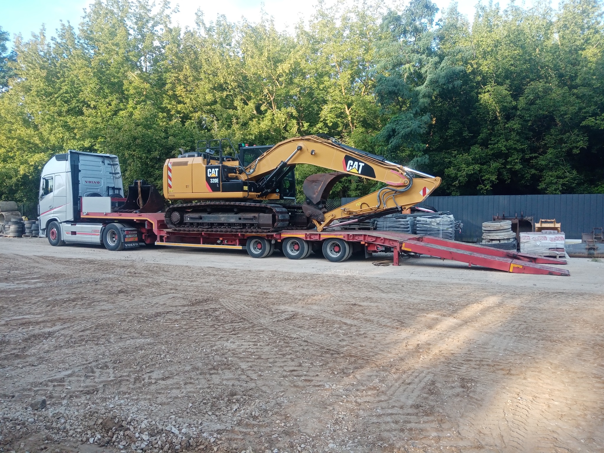 Ciężarówka Volvo z naczepą niskopodwoziową przewożąca żółtą koparkę CAT 320E na tle zielonych drzew. Transport maszyny budowlanej.