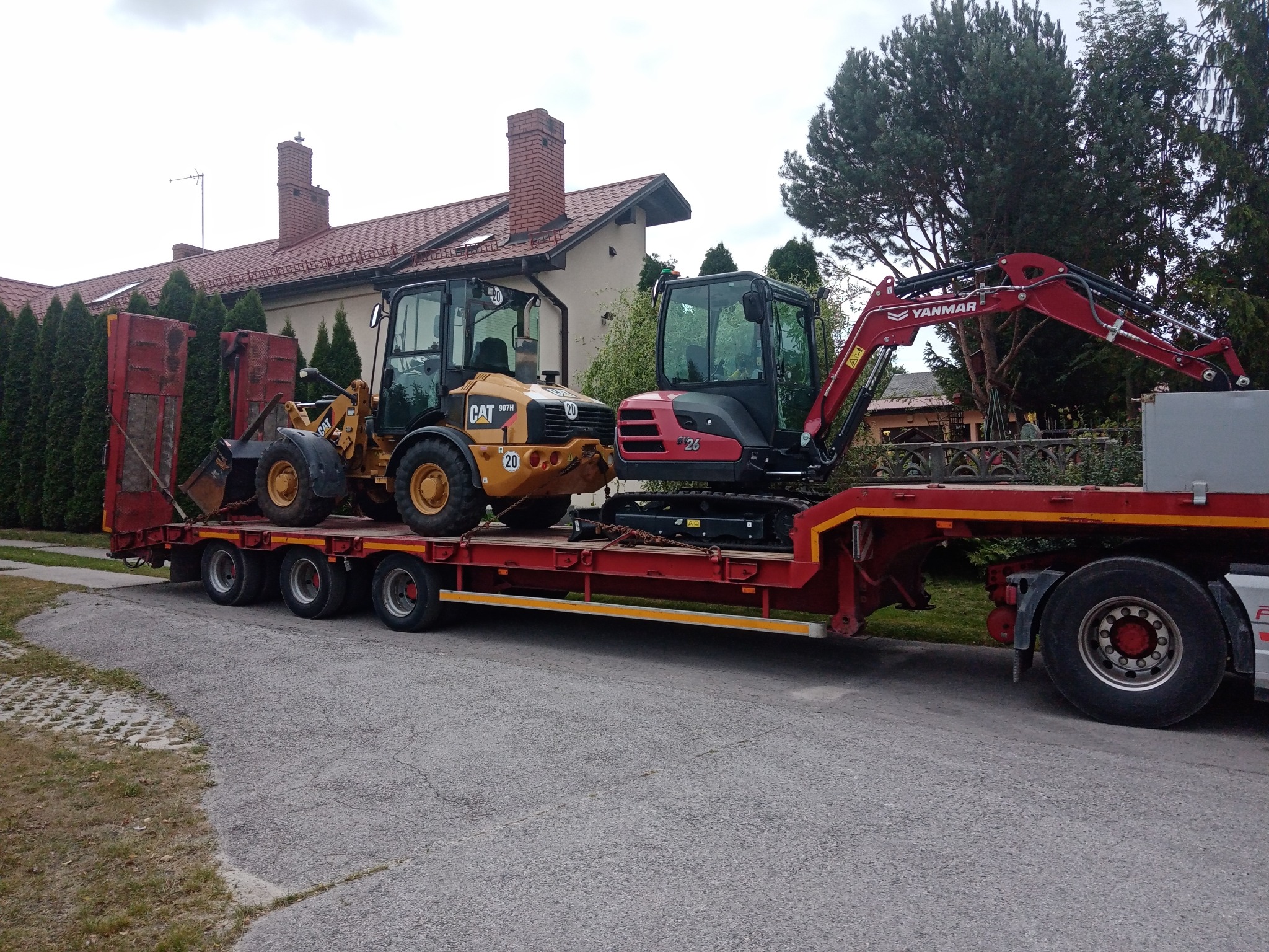 Laweta transportuje żółtą ładowarkę CAT i czerwoną koparkę YANMAR na czerwonej platformie, na tle domu z czerwonym dachem i zieleni.