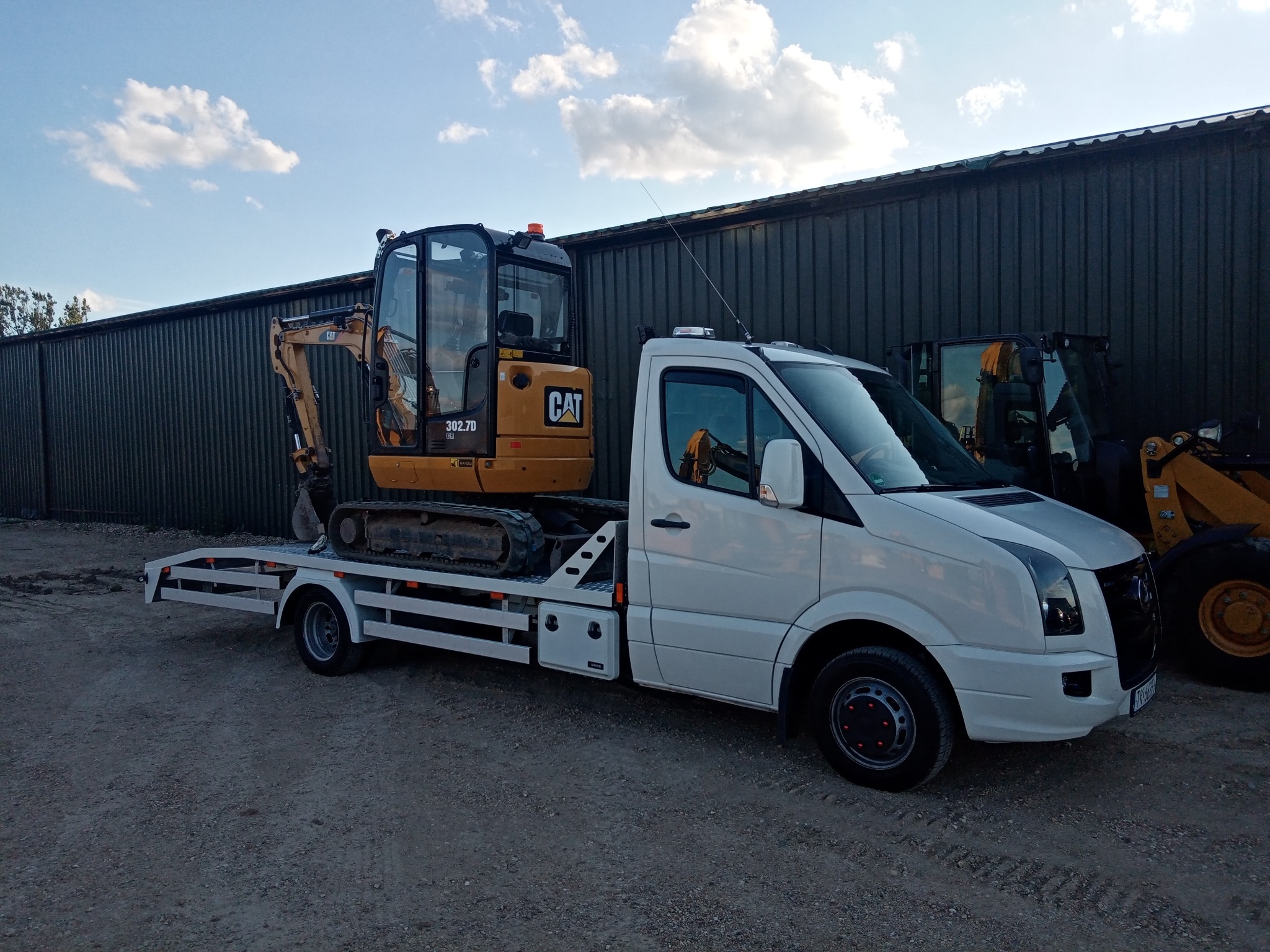 Biała laweta z żółtą mini koparką CAT 302.7D na platformie, zaparkowana na żwirowym placu przed szarozielonym budynkiem. Widoczny również fragment innej maszyny budowlanej.