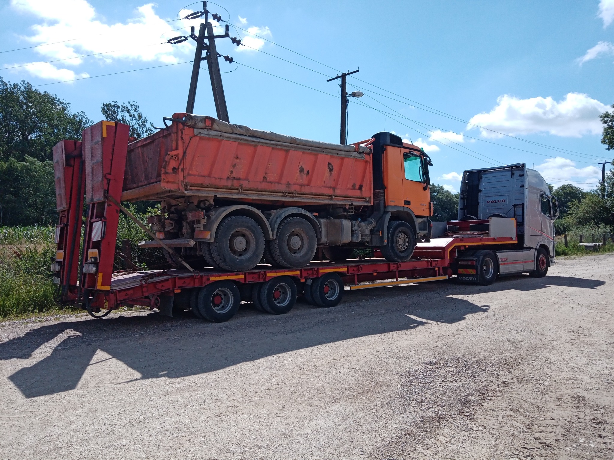 Pomarańczowa wywrotka na czerwonej platformie niskopodwoziowej, transportowana przez szary ciągnik Volvo, na żwirowej drodze w słoneczny dzień.