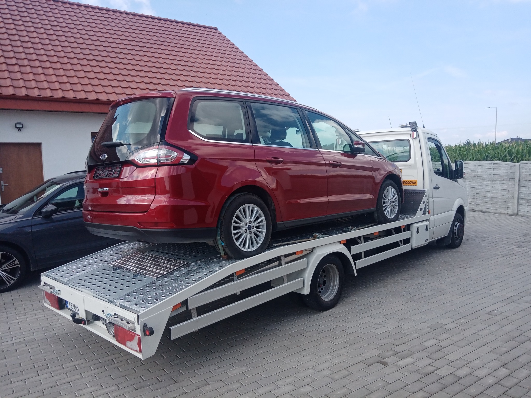 Czerwony Ford Galaxy na białej lawecie przed domem z czerwoną dachówką. Widoczny fragment zaparkowanego auta osobowego i betonowe ogrodzenie.
