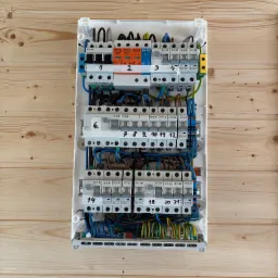 Rozdzielnica elektryczna z numerowanymi bezpiecznikami zamontowana na ścianie z drewnianych paneli. Widoczne przewody i elementy zabezpieczające.