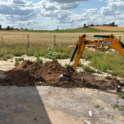 MAK-ELEKTRO usługi elektryczne - Żółta mini koparka JCB 15Z-1, kopie ziemię na tle pola i błękitnego nieba z chmurami. W tle widoczny budynek z drewnianą elewacją i narzędzia.