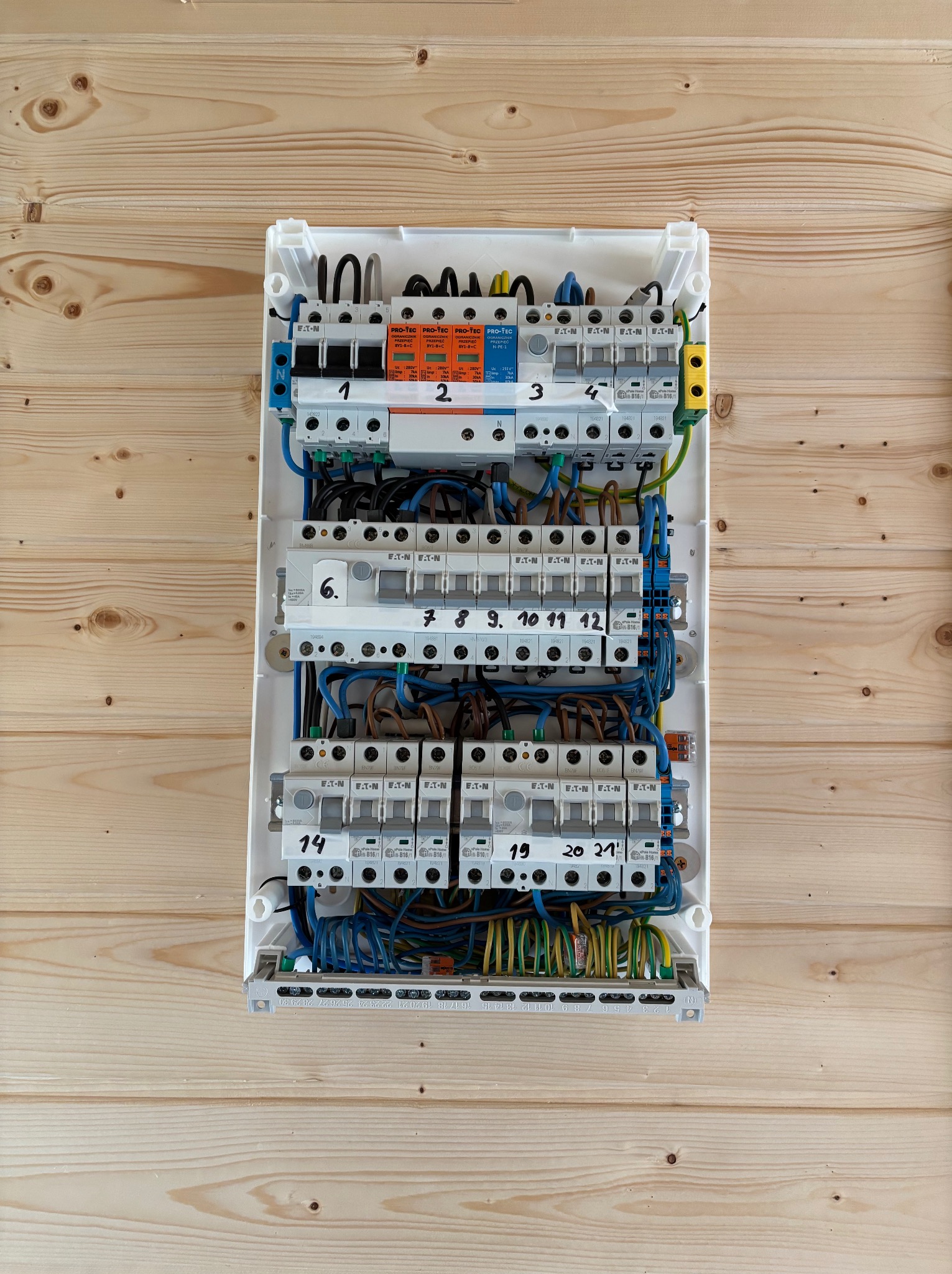 Rozdzielnica elektryczna z numerowanymi bezpiecznikami zamontowana na ścianie z drewnianych paneli. Widoczne przewody i elementy zabezpieczające.