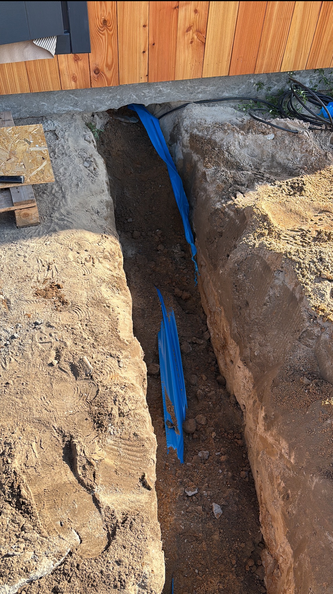 Wykop z niebieską rurą kablową ułożoną w ziemi przy drewnianej elewacji budynku. Widoczne ślady stóp na ziemi.