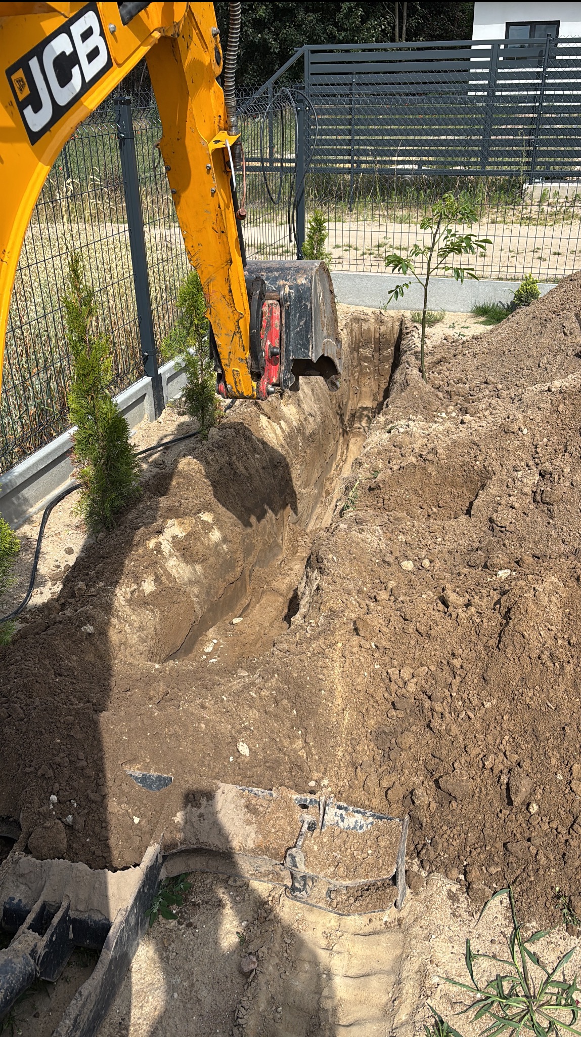 Żółta koparka JCB kopie wąski wykop w ziemi wzdłuż ogrodzenia z siatki. Świeżo wykopana ziemia tworzy wał po obu stronach wykopu. Widoczne rośliny i fragment budynku.