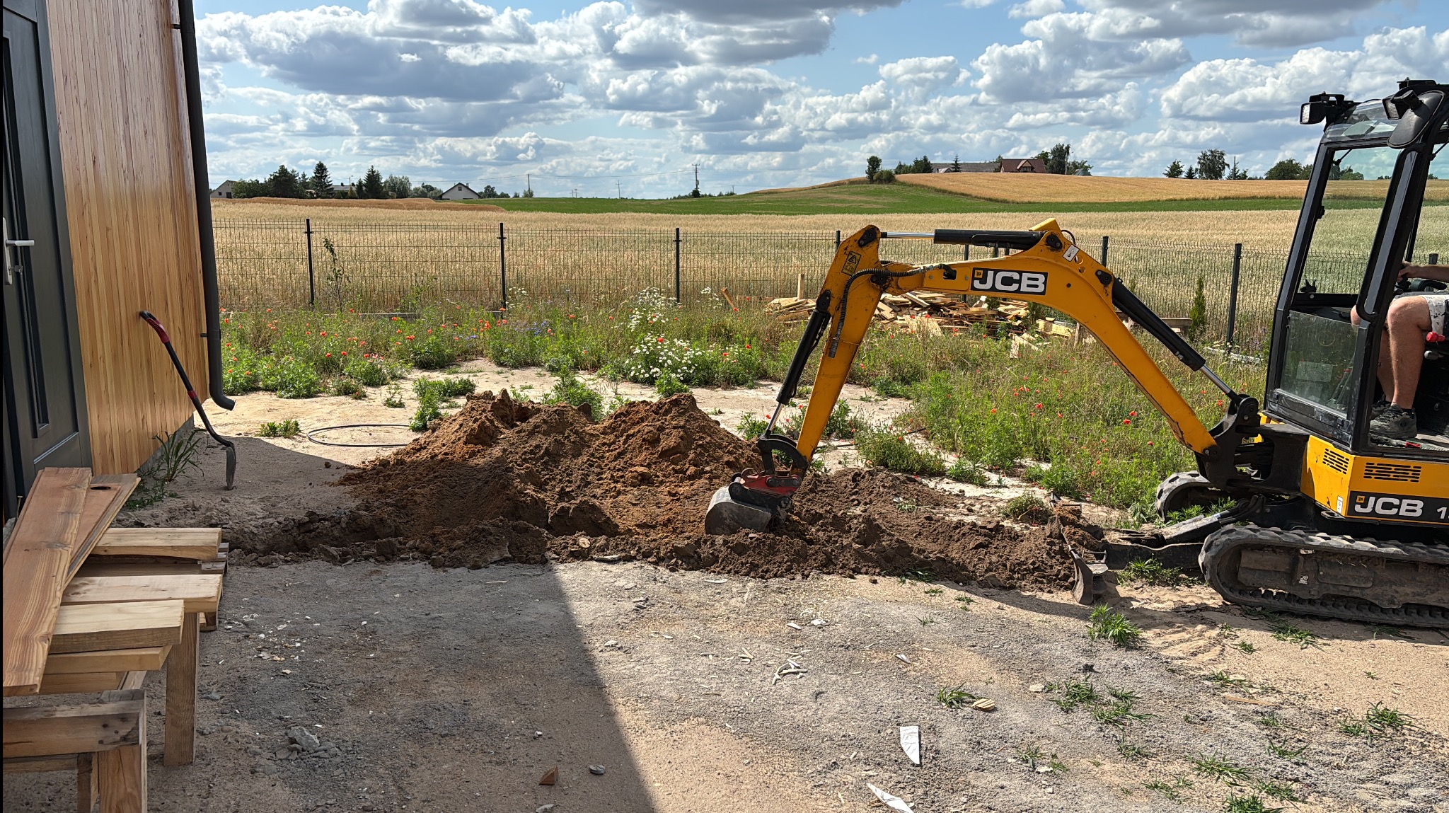 Żółta mini koparka JCB 15Z-1, kopie ziemię na tle pola i błękitnego nieba z chmurami. W tle widoczny budynek z drewnianą elewacją i narzędzia.