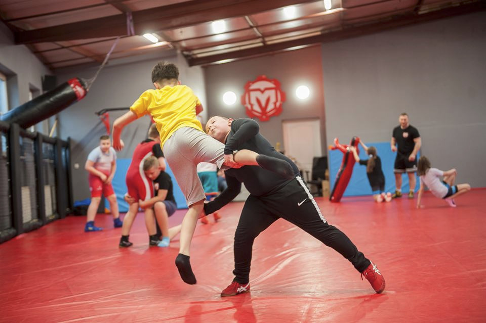 Trening sportów walki dla dzieci: chłopak w żółtej koszulce kopie drugiego w nogę podczas sparingu na czerwonej macie, w tle grupa ćwiczących pod okiem trenera.