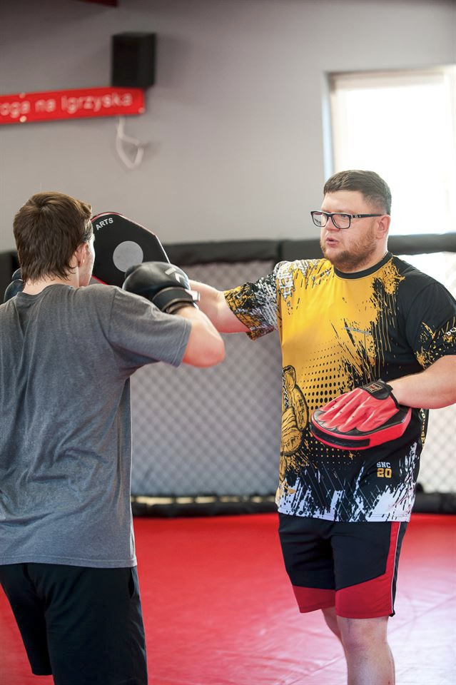 Trening sportów walki: mężczyzna z brodą w okularach, w koszulce z grafiką, trzyma czerwone rękawice bokserskie, podczas gdy drugi mężczyzna uderza w tarczę bokserską w czarnych rękawicach.