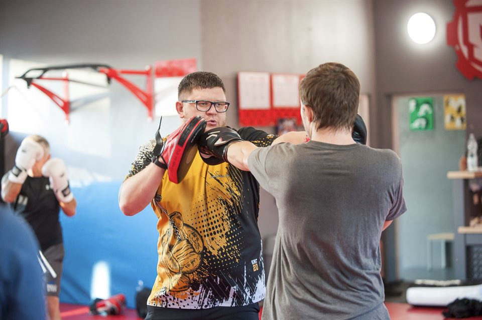 Trening sportów walki: mężczyzna w okularach z tarczami bokserskimi przyjmuje cios od partnera w szarej koszulce, w tle sala treningowa z wyposażeniem i innymi ćwiczącymi.