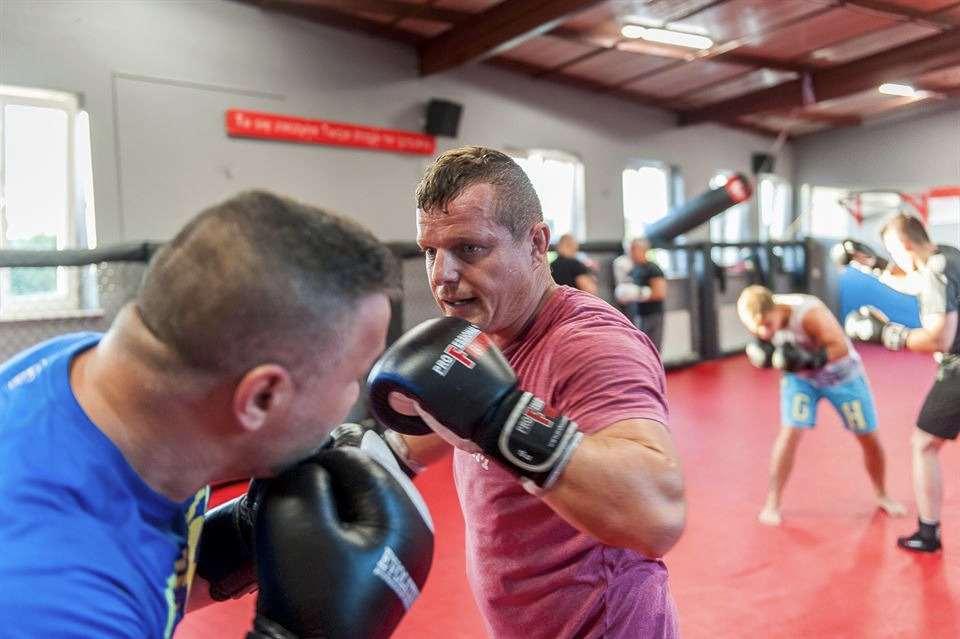 Dwóch mężczyzn w rękawicach bokserskich sparuje na czerwonej macie w sali treningowej, w tle inni trenujący.