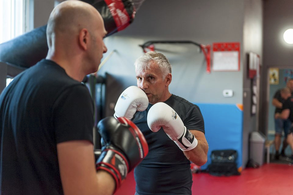 Trening boksu: mężczyzna w białych rękawicach bokserskich sparuje z trenerem w czarnych rękawicach, skupiony wzrok, sala treningowa z workiem bokserskim w tle.