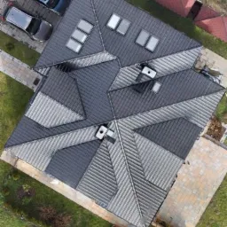 Widok z góry na dach domu z blachy trapezowej w odcieniach szarości z oknami dachowymi, dwa samochody zaparkowane obok.