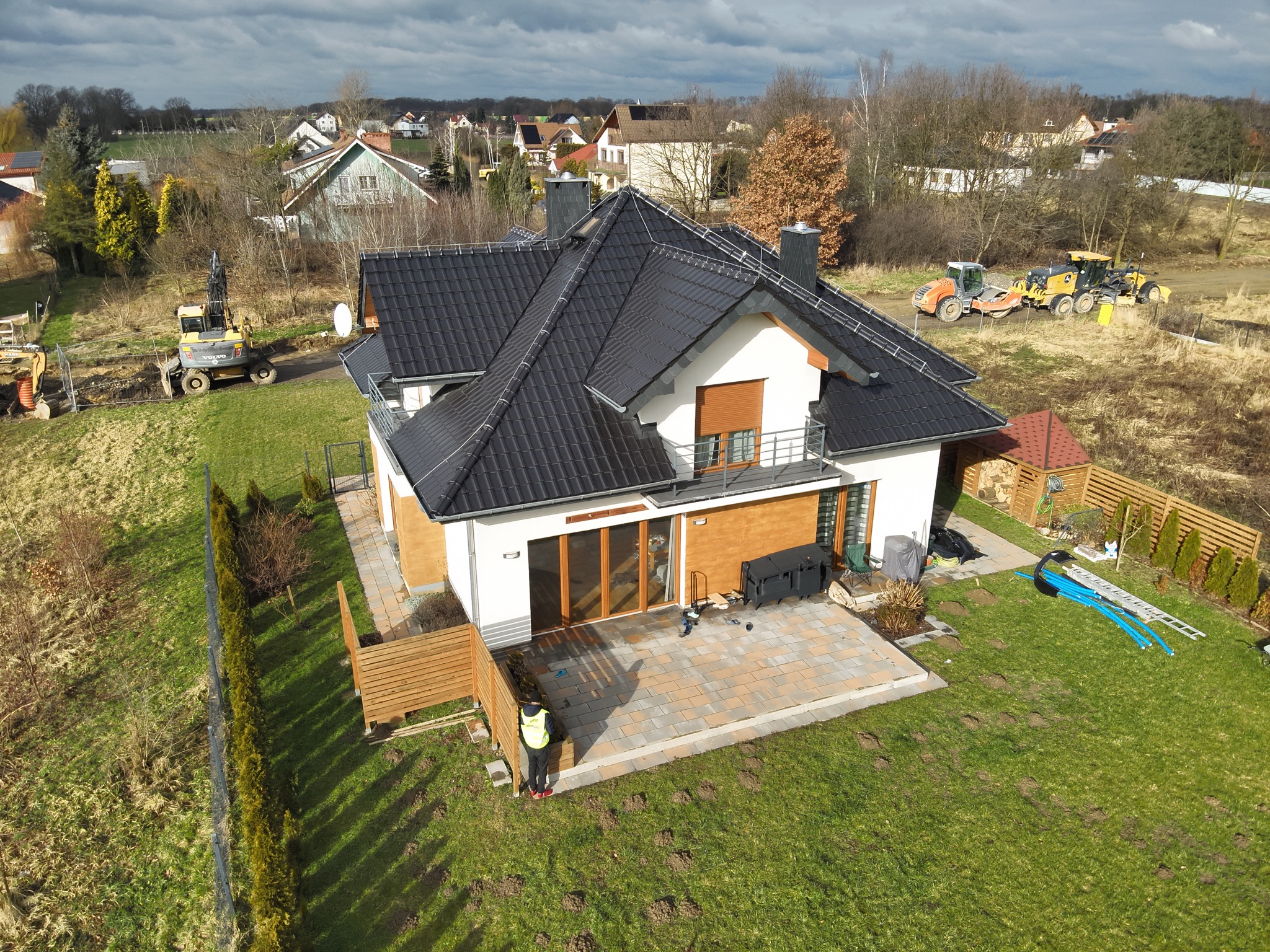 Widok z lotu ptaka na nowoczesny dom z ciemną dachówką, tarasem z kostki brukowej i zagospodarowanym ogrodem; w tle sprzęt budowlany i inne domy.