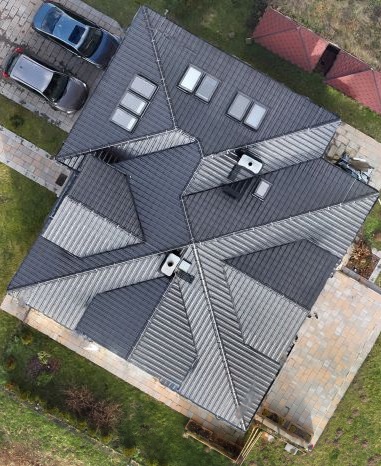 Widok z góry na dach domu z blachy trapezowej w odcieniach szarości z oknami dachowymi, dwa samochody zaparkowane obok.