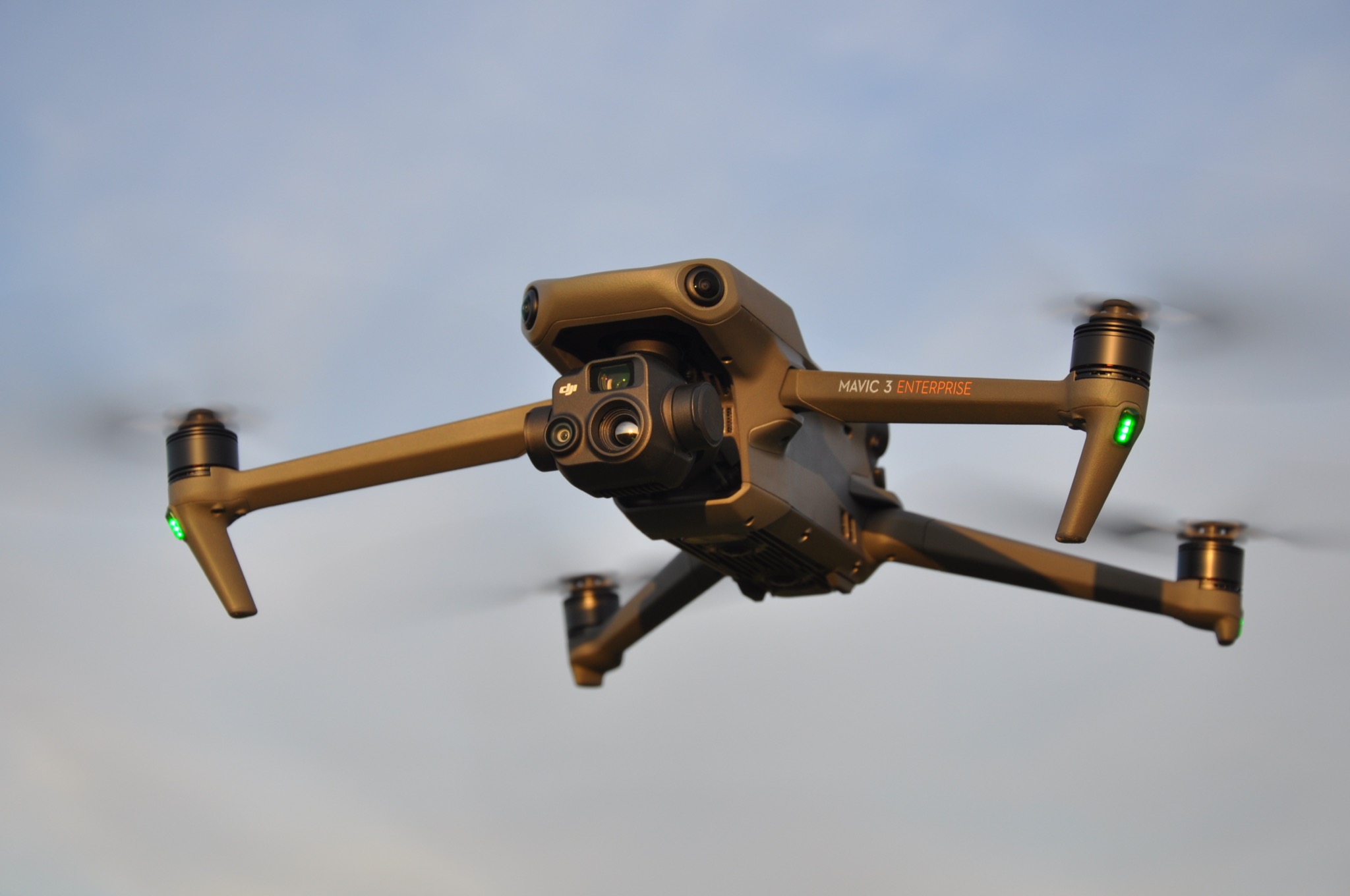 Latający dron Mavic 3 Enterprise z włączonymi zielonymi diodami LED na ramionach, na tle jasnego nieba.