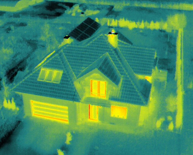 Termowizyjne zdjęcie domu jednorodzinnego z panelami słonecznymi na dachu, ukazujące straty ciepła przez okna i bramę garażową.