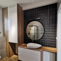 Nowoczesna łazienka z czarnymi, trójwymiarowymi płytkami, okrągłym lustrem odbijającym kabinę prysznicową, białą umywalką nablatową, szafką z drewnianym blatem i podłogą imitującą drewno.
