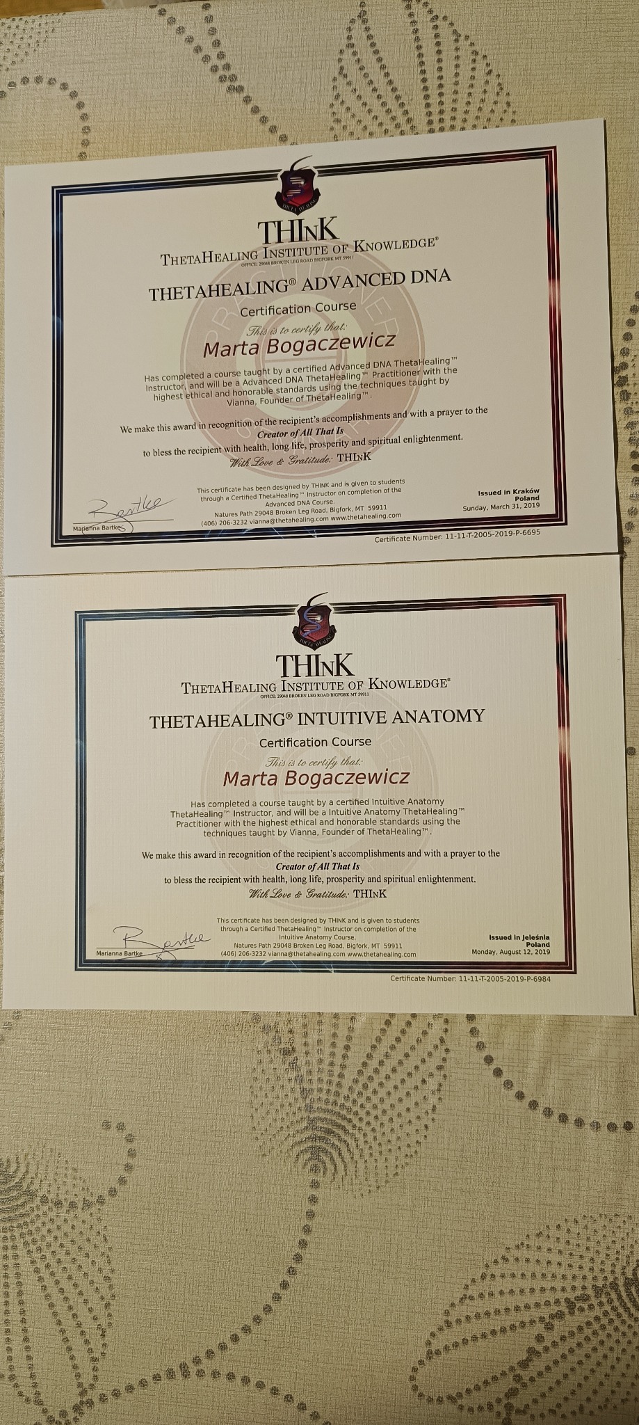 Dwa certyfikaty ThetaHealing Institute of Knowledge dla Marty Bogaczewicz, jeden z kursu Advanced DNA, drugi z Intuitive Anatomy, oba oprawione w ramki z bordową obwódką, leżące na beżowej...