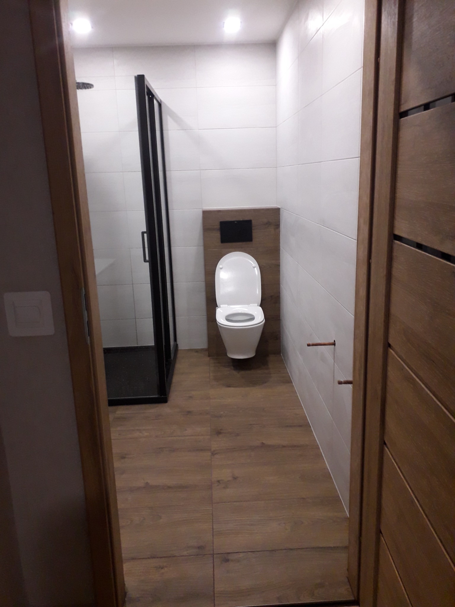 Nowoczesna łazienka z prysznicem typu walk-in, białymi płytkami i drewnopodobną podłogą, widziana zza drewnianej ościeżnicy.