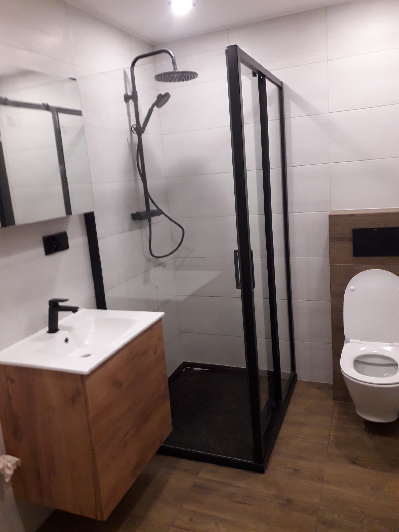 Nowoczesna łazienka z kabiną prysznicową z czarną ramą, umywalką z szafką w kolorze drewna i toaletą.