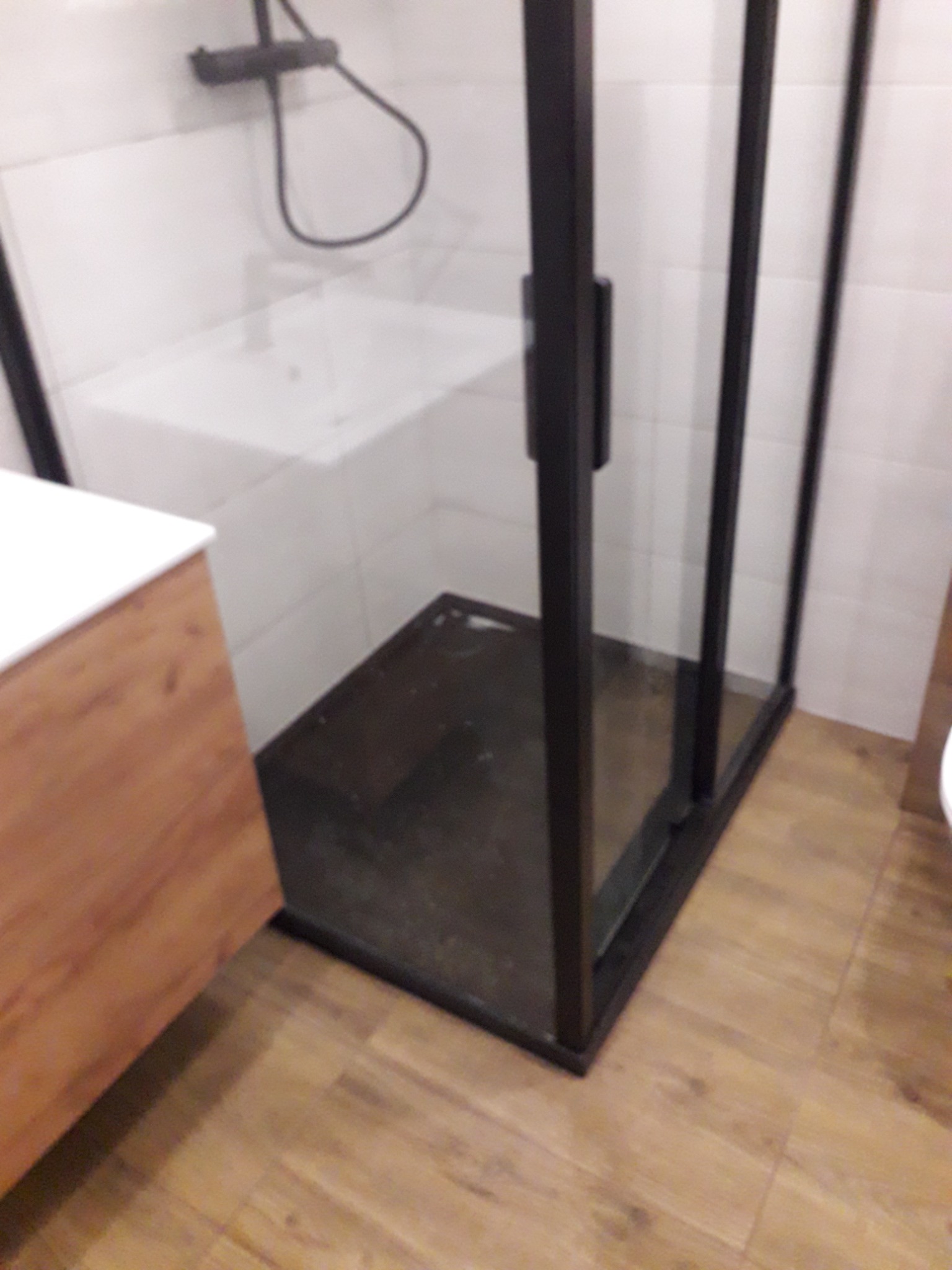 Nowoczesna kabina prysznicowa z czarnymi ramami, widok na armaturę i umywalkę w tle, drewnopodobna podłoga.