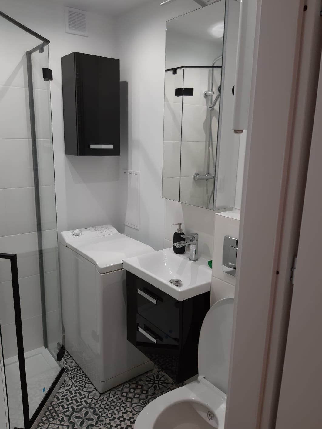 Funkcjonalna, mała łazienka z kabiną prysznicową, pralką, umywalką z czarną szafką i toaletą, wykończona białymi płytkami i wzorzystą podłogą.