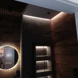 Nowoczesna łazienka z drewnopodobnymi płytkami, kamienną ścianą z podświetlanymi półkami, okrągłym lustrem z podświetleniem LED i deszczownicą.