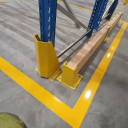 Oznakowanie poziome żółtą farbą w magazynie, zabezpieczenie narożnika regału magazynowego żółtym odbojnikiem, drewniana belka ochronna przy podstawie regału.
