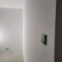 Przygotowanie do malowania ścian: gładka, biała ściana z zabezpieczonym gniazdkiem elektrycznym zieloną taśmą malarską, surowa podłoga z resztkami gładzi.