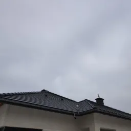 Nowoczesny, dwuspadowy dach pokryty czarną dachówką ceramiczną, z widocznymi rynnami i kominem na tle pochmurnego nieba.