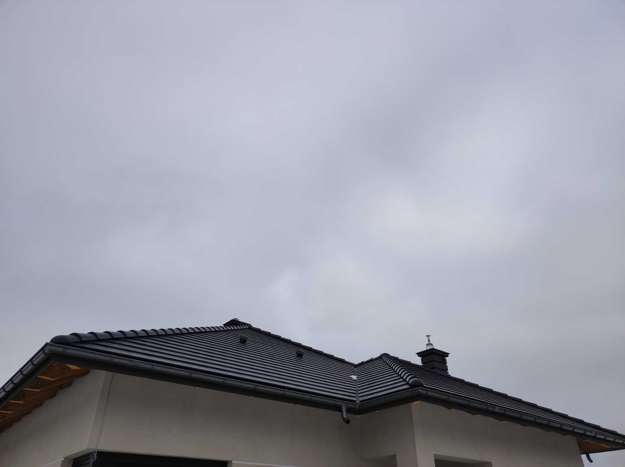 Nowoczesny, dwuspadowy dach pokryty czarną dachówką ceramiczną, z widocznymi rynnami i kominem na tle pochmurnego nieba.