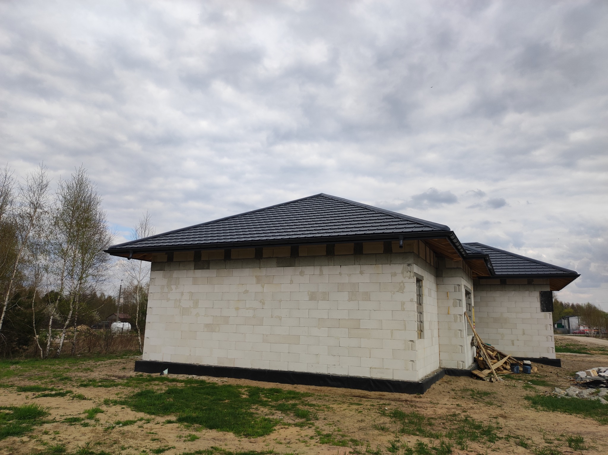 Surowy stan budynku z białych bloczków z zamontowanym czarnym dachem z dachówki ceramicznej i rynnami, widok na boczną ścianę i dach, na ziemi leżą drewniane elementy szalunku.