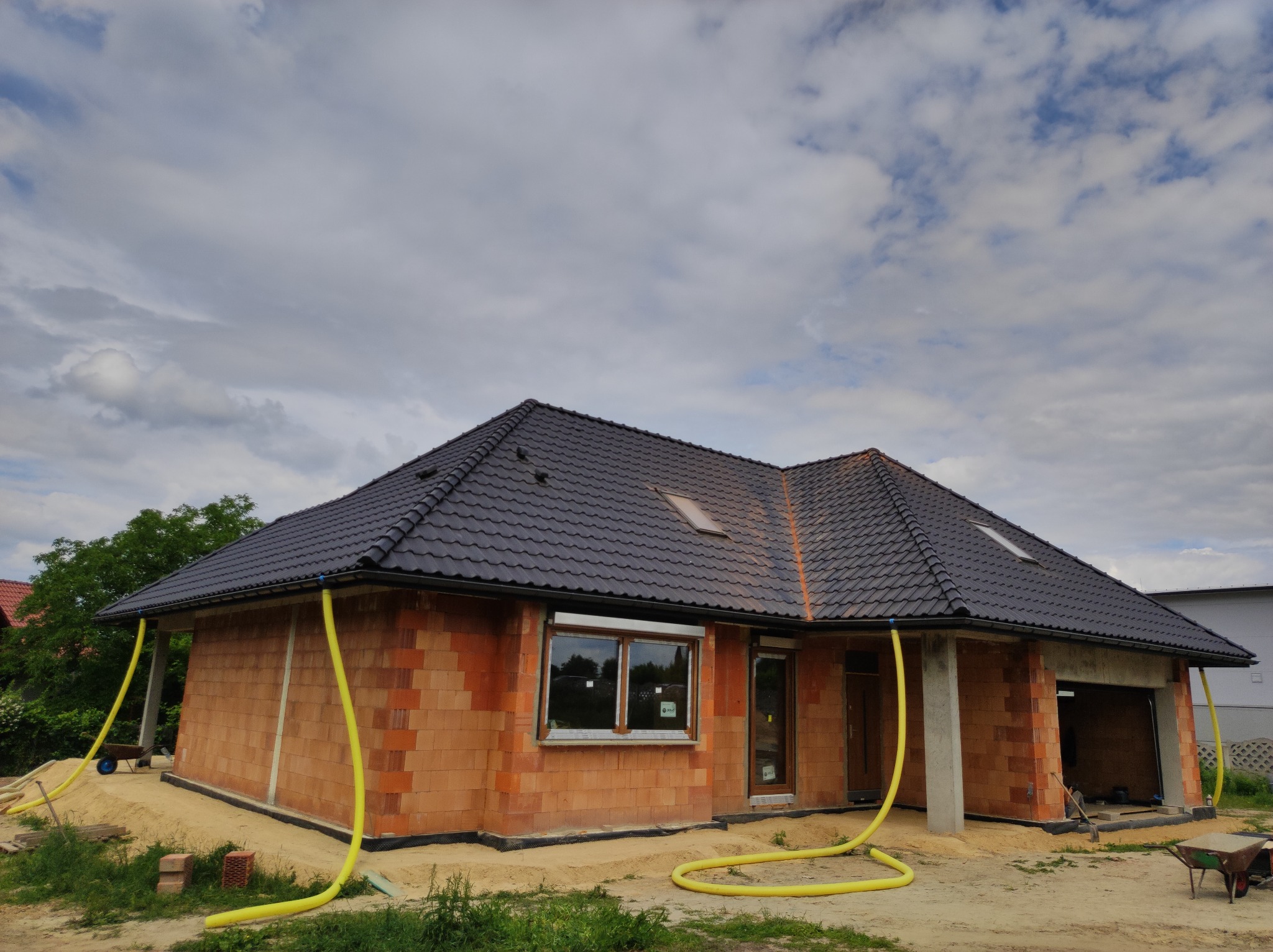 Dom w trakcie budowy z ciemną dachówką, widoczne rynny i żółte węże wentylacyjne na zewnątrz, częściowo otynkowane ściany z czerwonej cegły.